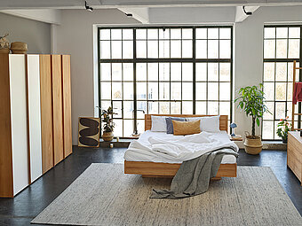 Ein modernes Schlafzimmer von Team 7 in einem Loft gezeigt, das Bett hat eine schwebende Optik und wirkt somit sehr leicht, auch in einem kleinen Schlafzimmer